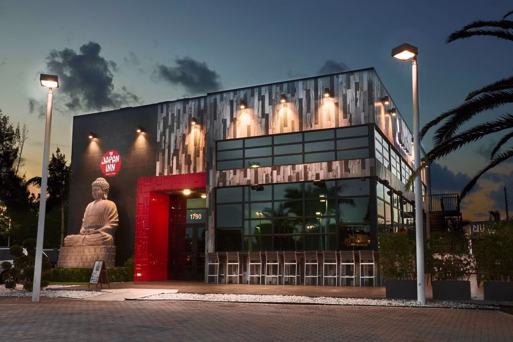 A contemporary restaurant exterior featuring a large red entrance and a decorative Buddha statue. The building has glass windows and modern architecture, set against a twilight sky. This venue offers fresh sushi rolls in South Florida.