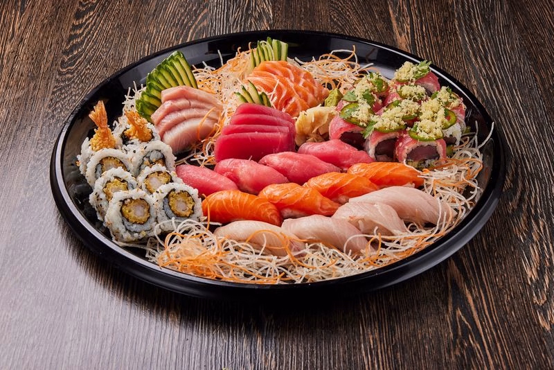 A circular black platter displays an assortment of sushi, including various types of sashimi and nigiri, decorated with shredded vegetables and garnishes. This vibrant presentation highlights options suitable for catering for International Soccer Season.