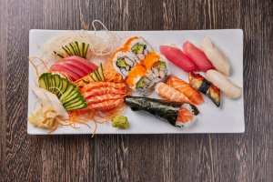 A white rectangular plate filled with various types of sushi, including sashimi slices, rolls, and nigiri. The assortment features salmon, tuna, and eel, along with garnishes of ginger and wasabi, presented on a wooden surface. Ideal for teppanyaki dining in Plantation.