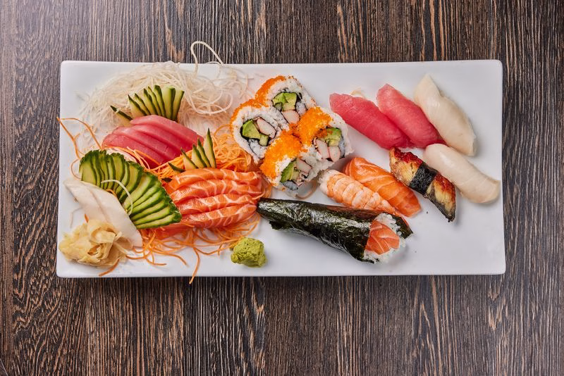A white rectangular plate filled with various types of sushi, including sashimi slices, rolls, and nigiri. The assortment features salmon, tuna, and eel, along with garnishes of ginger and wasabi, presented on a wooden surface. Ideal for teppanyaki dining in Plantation.