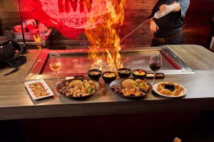 A table at a Japanese restaurant in Plantation features four dishes including chicken, shrimp, vegetables, fried rice, and sushi rolls. Flames erupt from a grill beside the dishes while drinks are placed on the table.