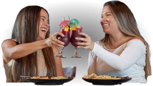 Two women with long hair hold up red drinks in stemmed glasses while seated at a table with plates of food against a black background.