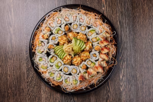 A large sushi platter with assorted maki and nigiri arranged in a circle on a black tray, topped with sliced avocado and served on a wooden table.