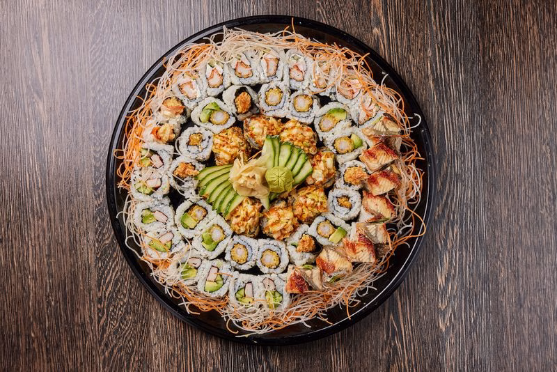 A large sushi platter with assorted maki and nigiri arranged in a circle on a black tray, topped with sliced avocado and served on a wooden table.