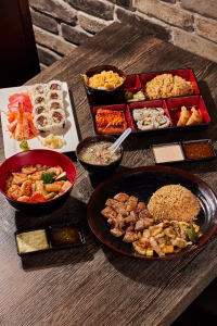 A table spread of assorted Japanese dishes, including sushi, rice bowls, and grilled meat, arranged in black and red serving dishes on a wooden tabletop.