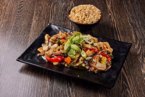 A black rectangular plate of stir-fried noodles with vegetables and herbs sits on a dark wooden table, with a bowl of rice behind it.