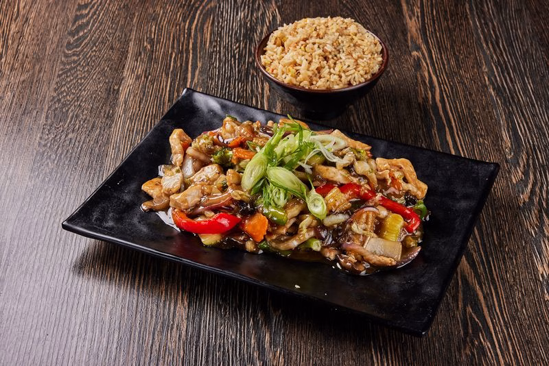A black rectangular plate of stir-fried noodles with vegetables and herbs sits on a dark wooden table, with a bowl of rice behind it.